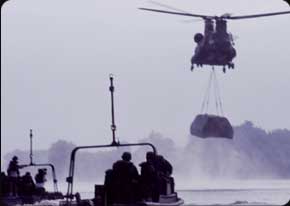 A Chinook sling-loads a bridge segment into position during a river crossing exercise.  Bridge Crewmembers (MOS 12C) await in boats to assemble many of these segments into an entire bridge span.